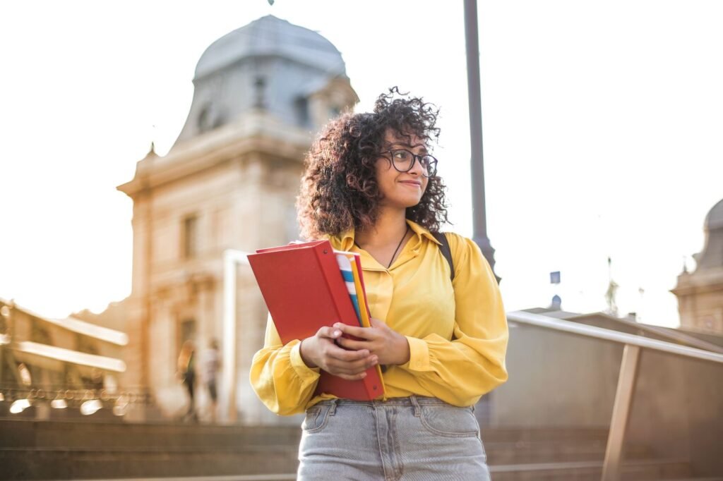A smiling student with curly hair and glasses holding books on campus steps