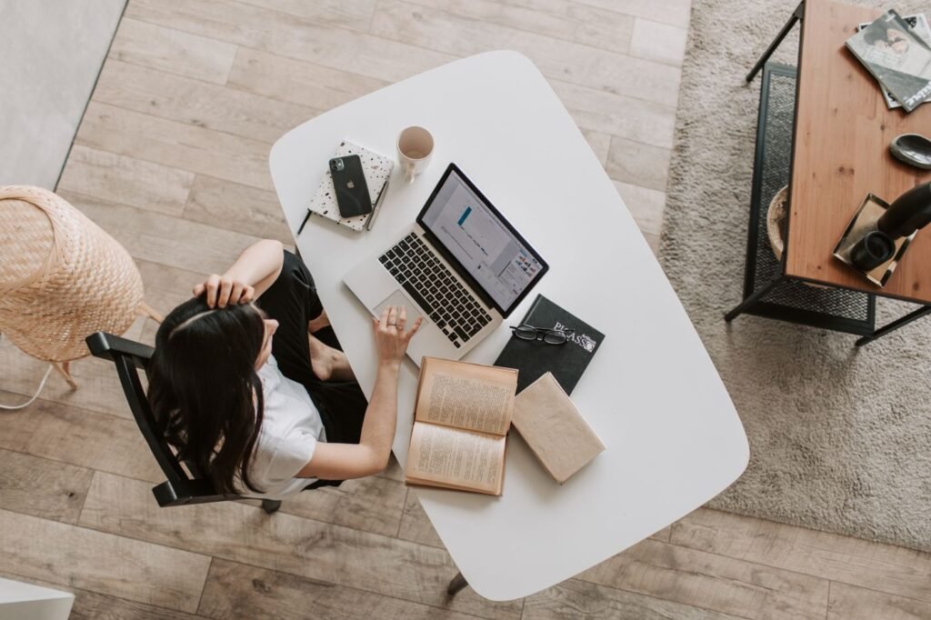 A young woman working with a laptop and an open book at a home desk