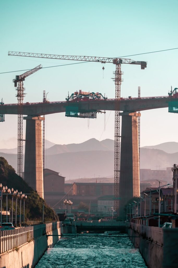 A large bridge under construction at sunrise, with cranes and mountains behind